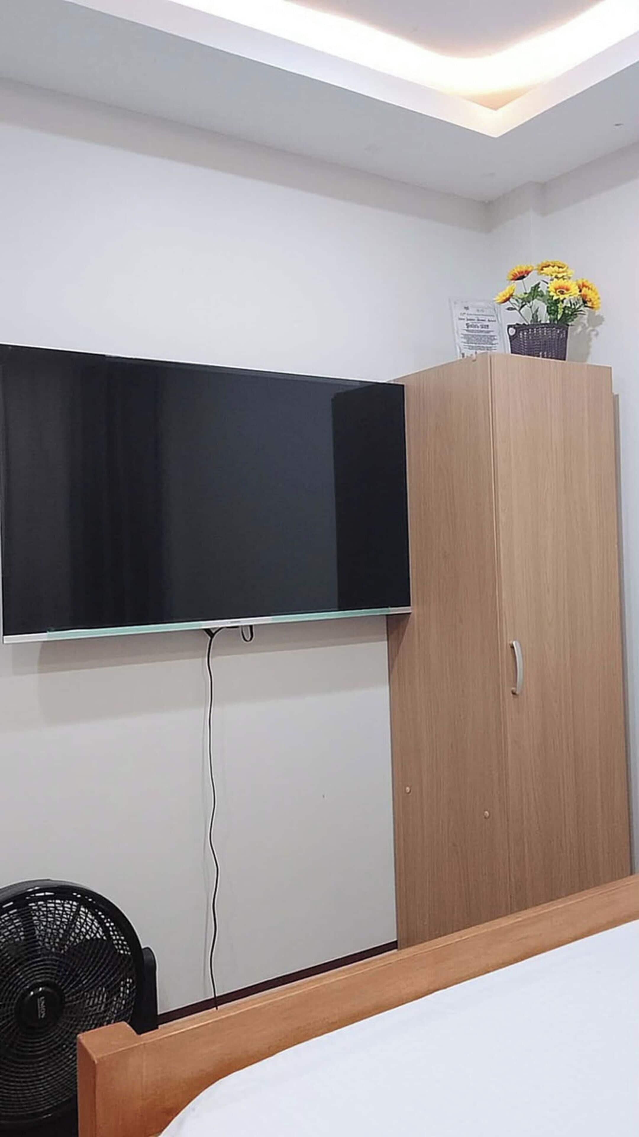 Een slaapkamer met een bed, een aan de muur bevestigde televisie, een houten kledingkast en een ventilator.