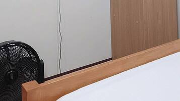 Een slaapkamer met een bed, een aan de muur bevestigde televisie, een houten kledingkast en een ventilator.