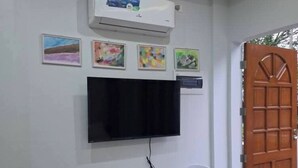 A room with a flat-screen TV mounted on the wall, two white plastic chairs, a small table, and a wooden floor. - Cozy Stay at JAL Transient House (Hinoba-an)