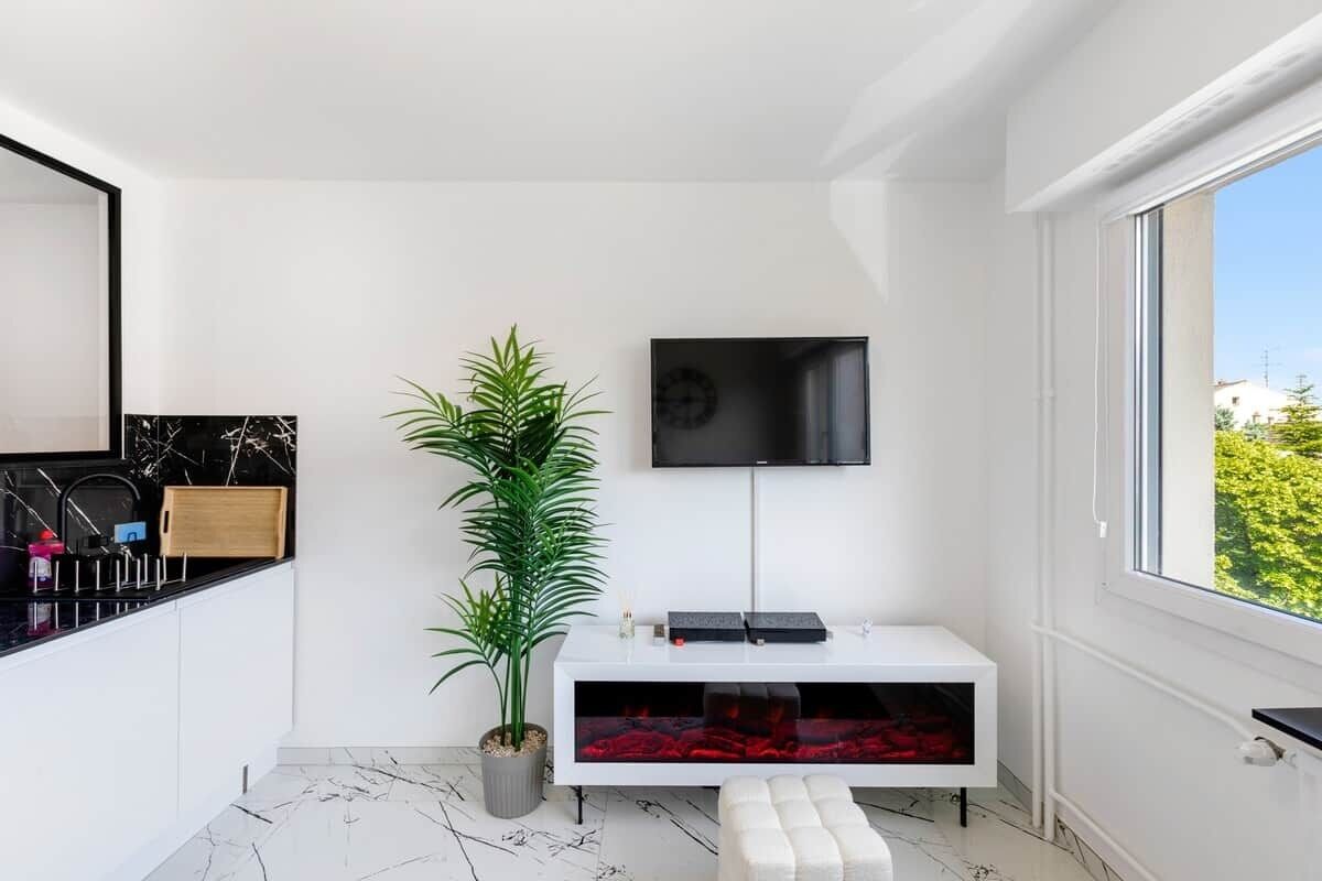 A modern living room with a white console table, a large potted plant, a flat-screen TV, and a window with a view of greenery.