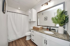 Un baño con inodoro blanco, cortina de ducha, lavabo con espejo y una planta en maceta.
