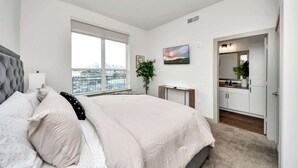 Um quarto com uma cama grande, uma janela com vista para a cidade, uma mesinha de cabeceira e uma pequena planta.