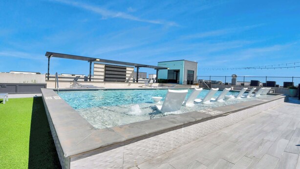 Uma piscina no terraço com céu azul límpido, área de estar coberta e um pequeno edifício ao fundo.