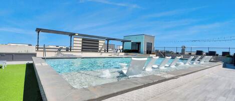 Une piscine sur le toit, avec un ciel bleu dégagé, un espace salon abrité et un petit bâtiment en arrière-plan.