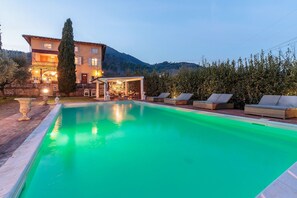 Ein Pool mit Liegestühlen, eine überdachte Sitzgelegenheit und ein Gebäude mit mehreren Fenstern und Lichtern.