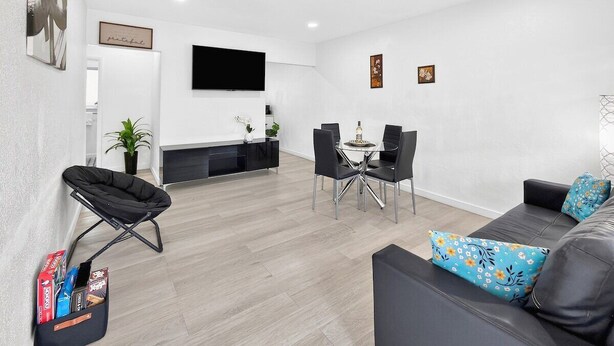 Una sala de estar moderna con un sofá, una mesa de comedor con sillas, un televisor de pantalla plana en la pared y una silla plegable negra.