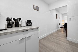 A modern kitchen with white cabinetry, a countertop with coffee maker and utensils, and a dining area with a chair and table. - Modern Miami Stay - Near Airport & Downtown (Miami)