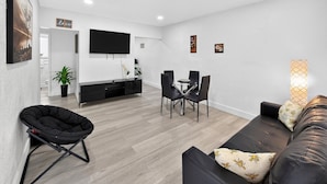 A modern living room with a sofa, a black chair, a dining table with chairs, a flat-screen TV mounted on the wall, and a potted plant. - Modern Miami Stay - Near Airport & Downtown (Miami)