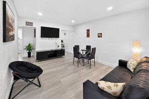A modern living room with a sofa, a black chair, a dining table with chairs, a flat-screen TV mounted on the wall, and a potted plant. - Modern Miami Stay - Near Airport & Downtown (Miami)