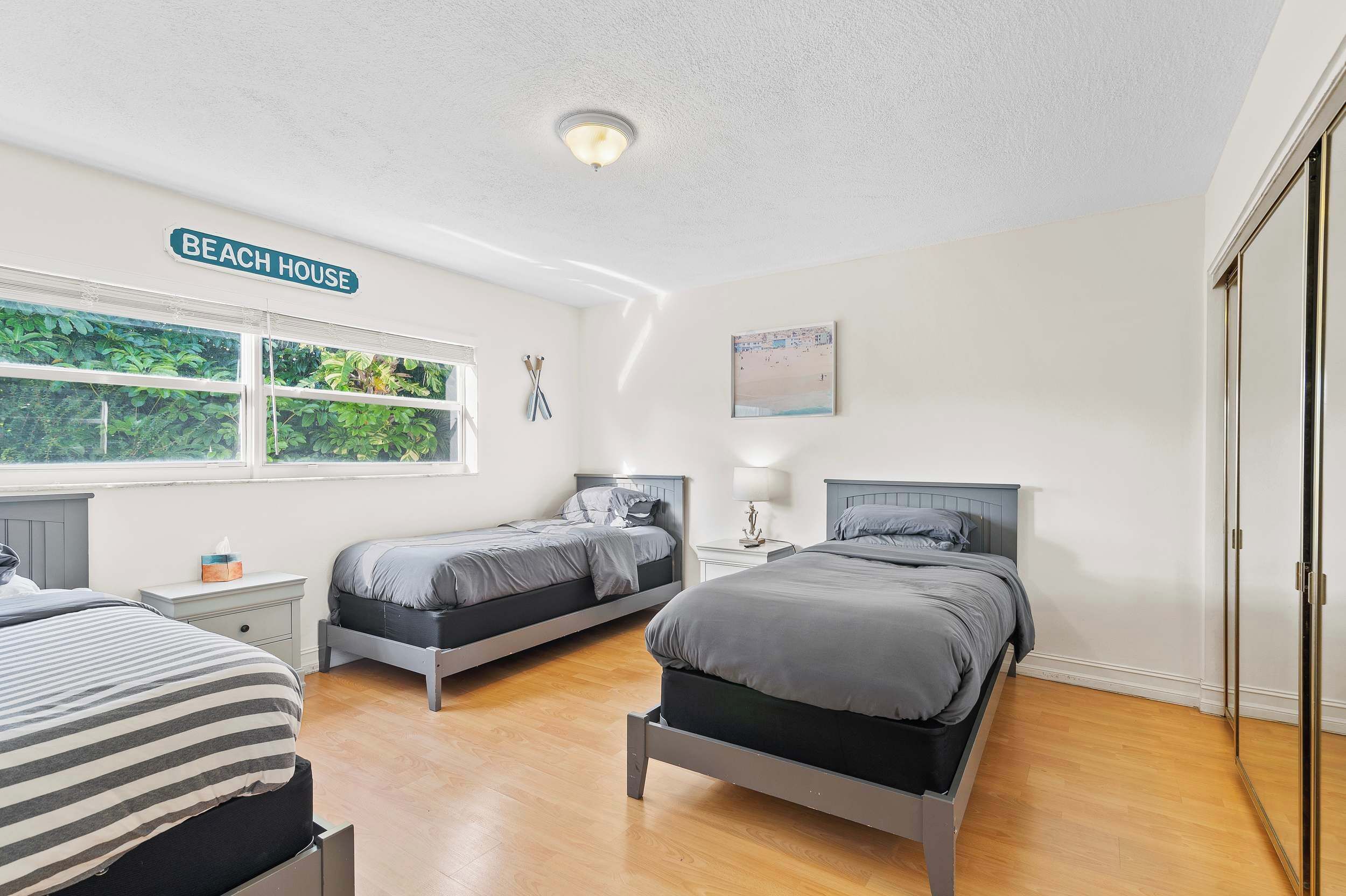 Ein Schlafzimmer mit zwei Betten, einem Fenster mit Blick auf Grünflächen, einer Deckenleuchte und einem Schild mit der Aufschrift 'Beach House'.