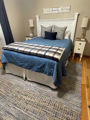 Un lit bien fait, recouvert d’une couvre-lit bleue et d’un plaid à motifs, flanqué de deux tables de chevet avec des lampes, le tout posé sur un tapis texturé et adossé à un mur orné d’un panneau indiquant « famille ».