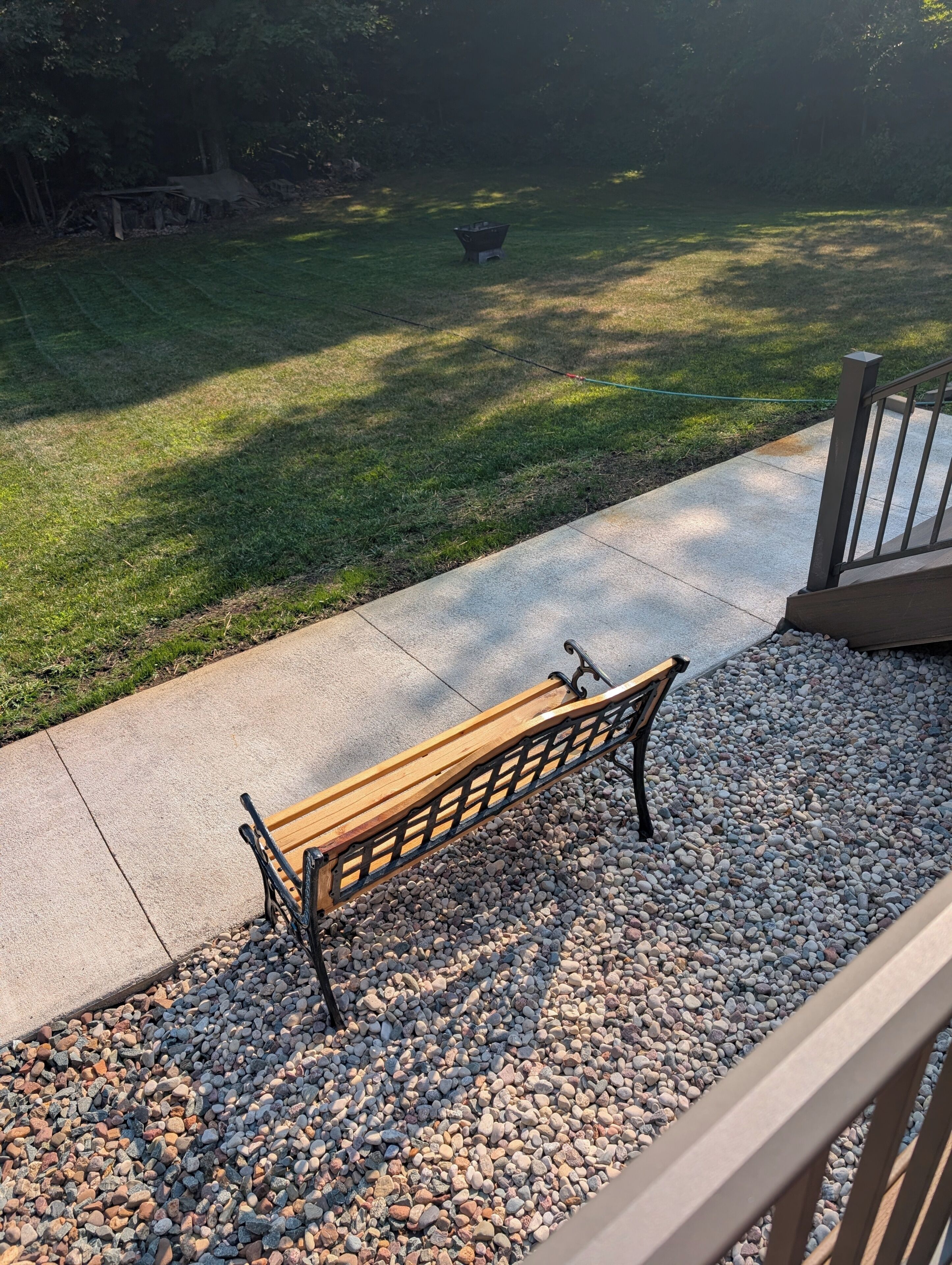A wooden bench on a concrete path with pebbles, leading to a grassy area with a wheelbarrow.