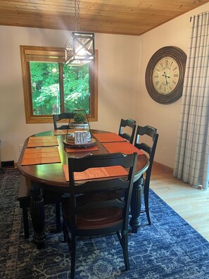 Un coin repas avec une table en bois, quatre chaises, une horloge au mur et une fenêtre donnant sur un paysage verdoyant.