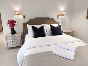Una cama bien hecha con sábanas blancas, dos mesitas de noche con lámparas y un jarrón con flores rosadas en una de ellas.
