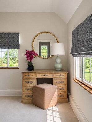 A room with a wooden dresser, a mirror with a gold bead garland, a lamp, and a window with a view of greenery.