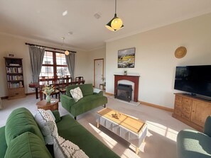 Ein Wohnzimmer mit grünem Sofa, einem Couchtisch, einem Kamin, einem Fernseher und einem Essbereich mit Tisch und Stühlen.