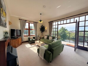 Ein geräumiges Wohnzimmer mit einem grünen Sofa, einem Couchtisch und einem Fernseher. Große Fenster mit Vorhängen, eine Pflanze und ein Blick ins Freie.