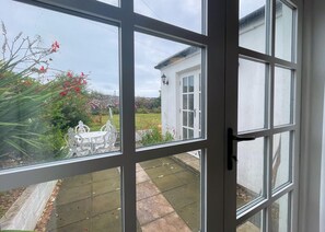 Blick durch eine Glastür mit weißer Außenfassade, einem Kiesweg und einem Garten mit Pflanzen, einem weißen Metalltisch und Stühlen.