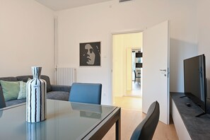 Ein modernes Wohnzimmer mit Glastisch, einer Couch, einem blauen Sessel, einer schwarzen Vase und einem Flachbildfernseher.