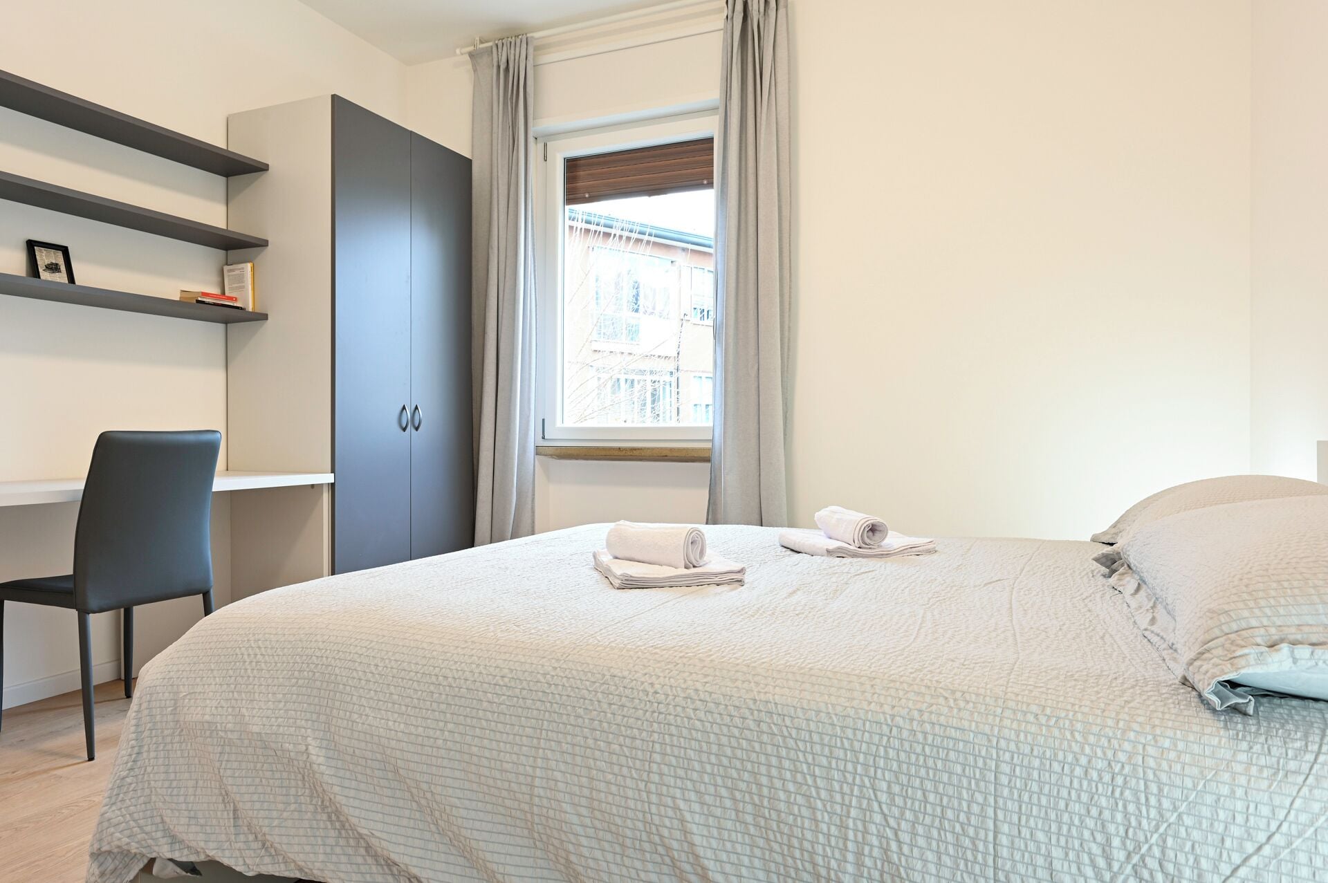 Une chambre à coucher avec un lit, un bureau et une chaise. Il y a une fenêtre avec des rideaux.
