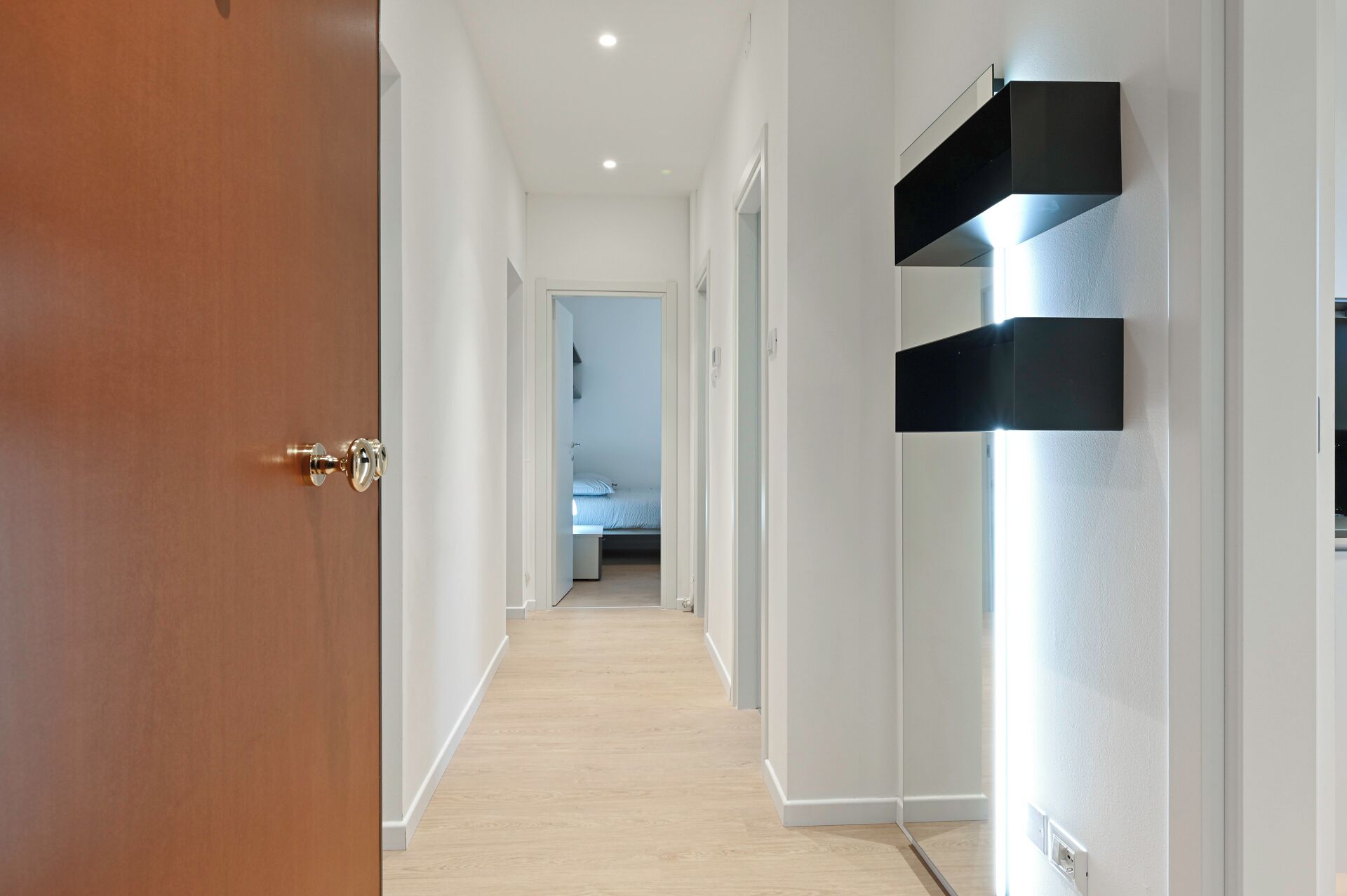 Un couloir avec une porte en bois, des murs blancs et un lit visible au loin.