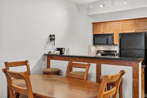 A compact kitchen with wooden cabinets, a black refrigerator, a microwave, and a coffee maker. There is a wooden dining table with two chairs. - The Suite Life (with AC unit!) by Cottonwood Lodging (Brighton)