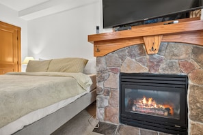 Ein Schlafzimmer mit Kamin aus Stein, einem Bett mit beiger Decke und einem Fernseher über dem Kamin.