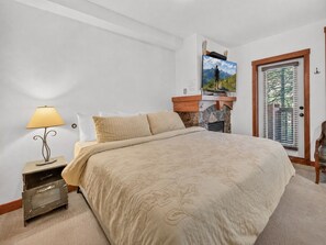 A bedroom with a bed, a nightstand, a lamp, a television, and a door with blinds. - The Suite Life (with AC unit!) by Cottonwood Lodging (Brighton)