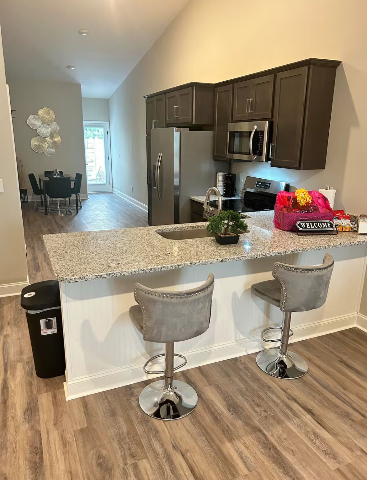 A modern kitchen with dark cabinets, stainless steel appliances, and a granite countertop. There are two bar stools and a trash bin.