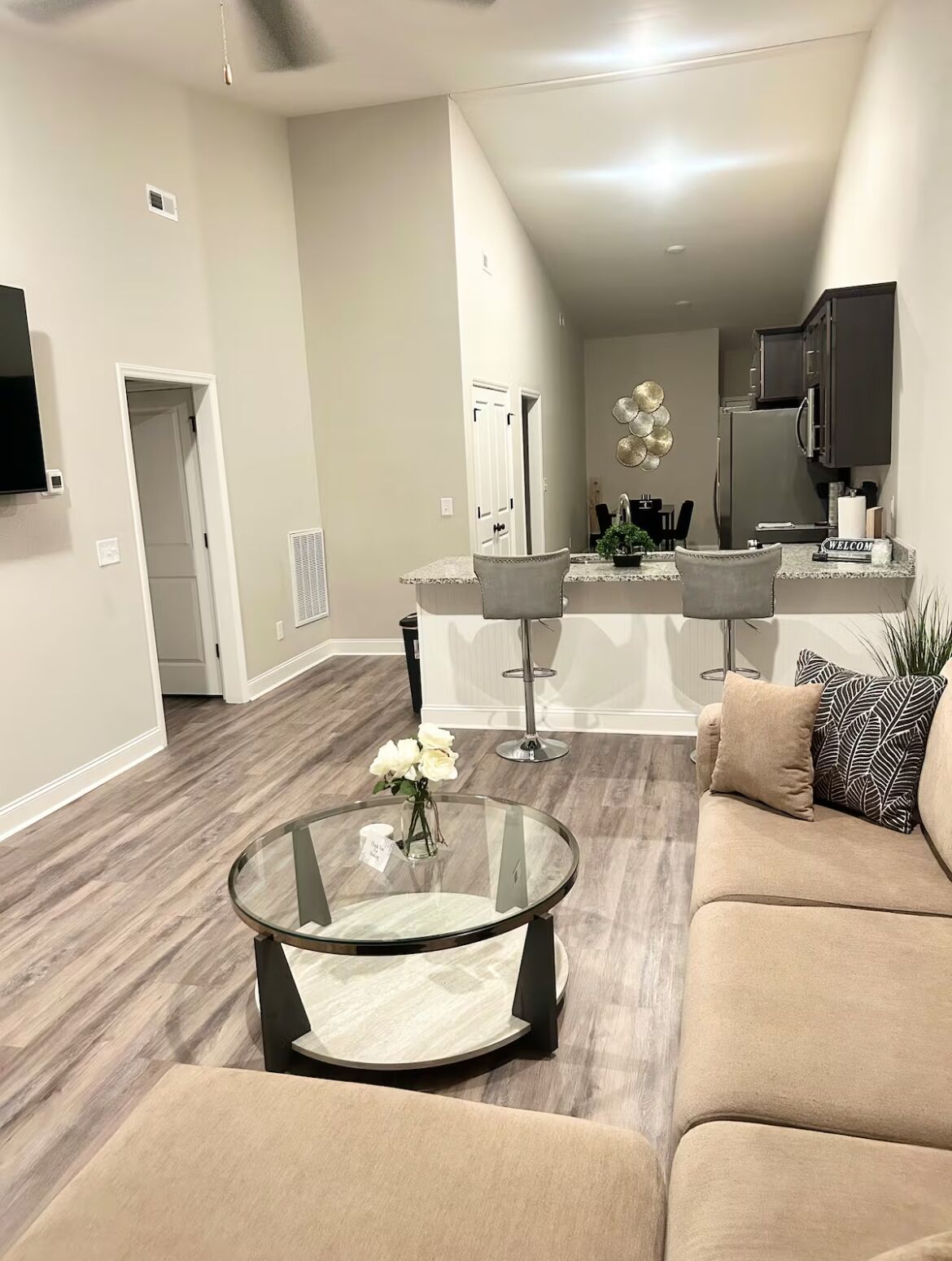 A modern living room with a glass coffee table, a beige sofa, and a kitchen area in the background.