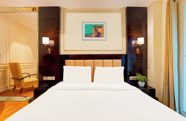 A hotel room with a large bed, two bedside lamps, a framed picture on the wall, a chair, and a potted plant.
