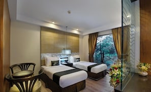 A hotel room with two beds, a sitting area with a chair, a small table, a nightstand, a lamp, and a view of trees through the window.