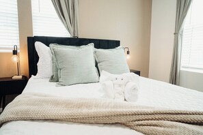 Un lit bien fait, avec une couette blanche et plusieurs oreillers, dont un ours en peluche blanc.