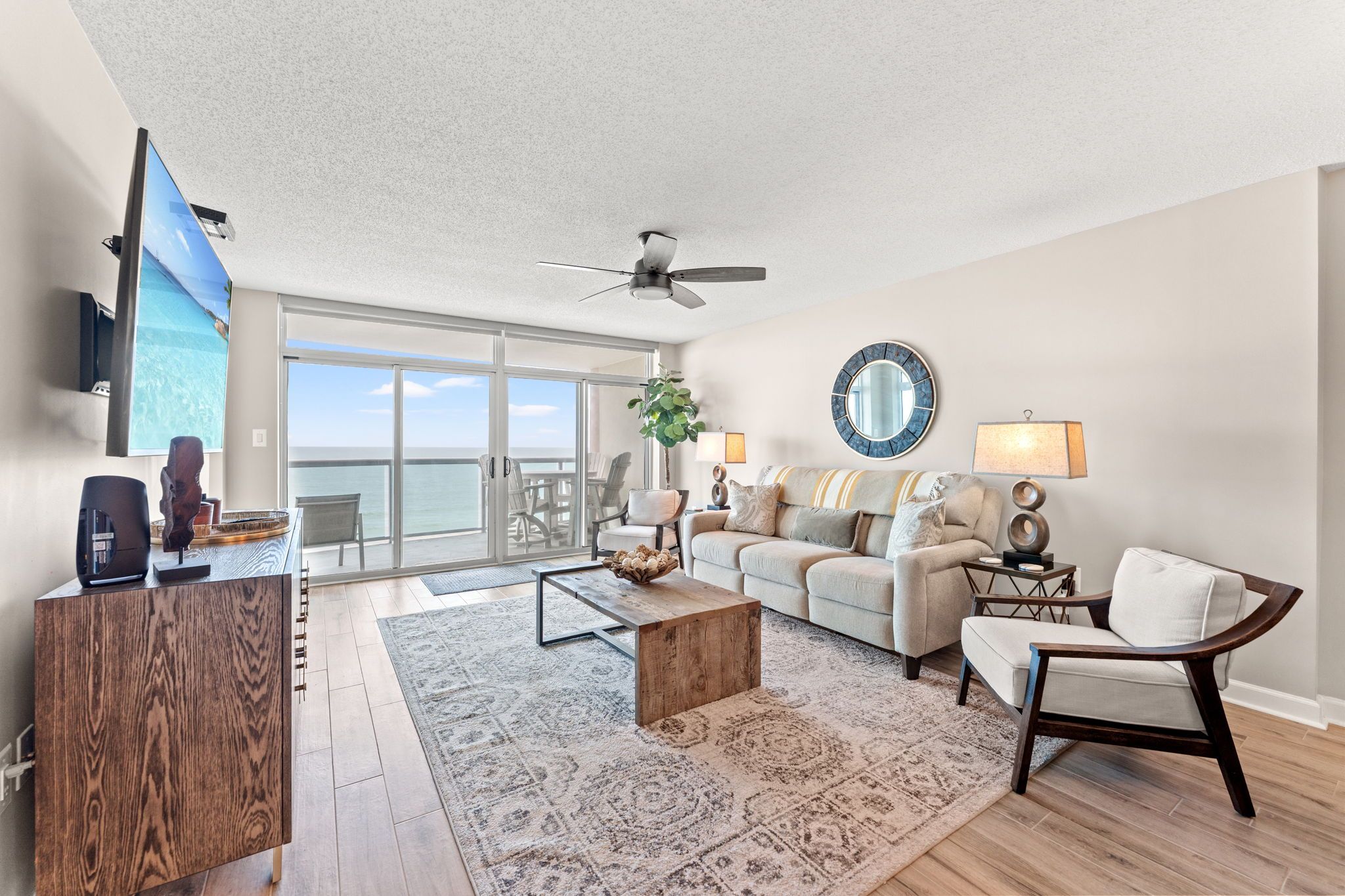 A spacious living room with a large window, a ceiling fan, a television, and a dining area with a view of the ocean.