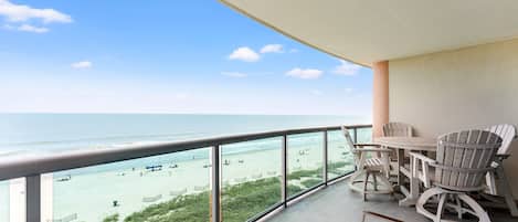 A balcony with a view of the beach, a table, and two chairs.