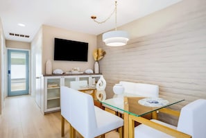 Ein moderner Essbereich mit einem Glastisch, weißen Stühlen, einem an der Wand befestigten Flachbildfernseher und einer Holzvitrine mit Dekorationsgegenständen.