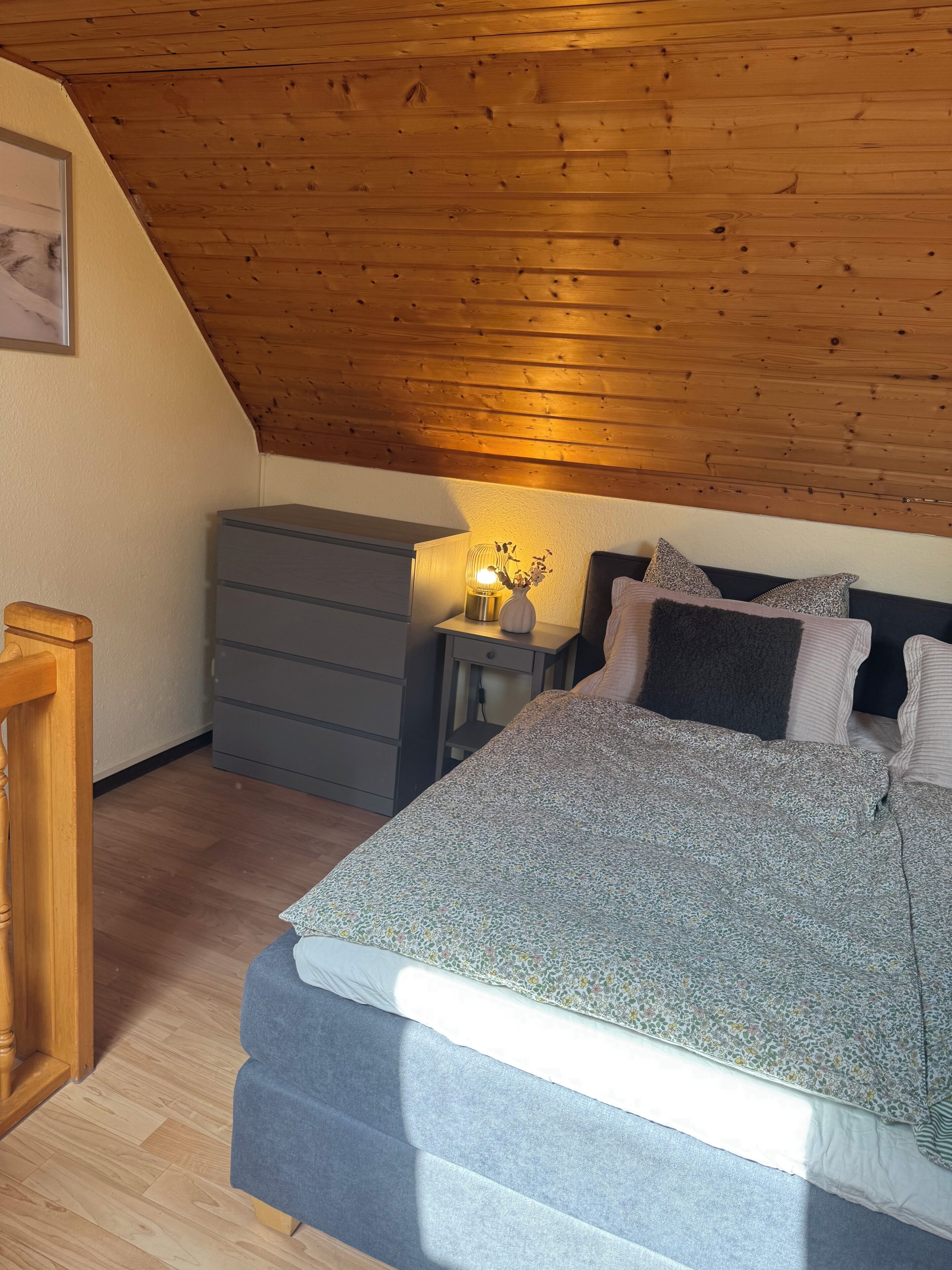 Een slaapkamer met schuine balken, een bed met beddengoed met patroon, een nachtkastje met lamp en een dressoir.