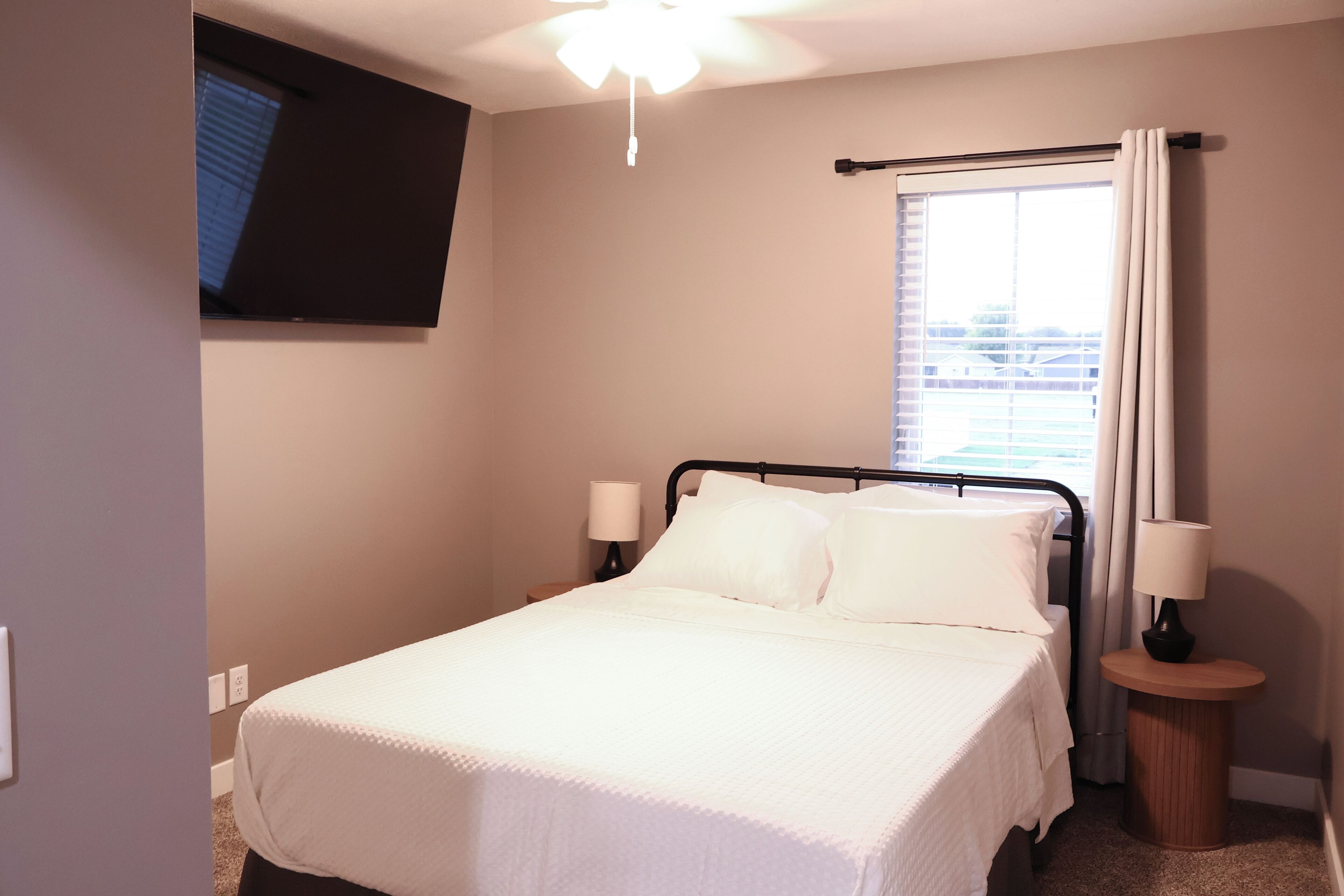 A hotel room with a bed, two bedside lamps, a window with curtains, and a ceiling fan.