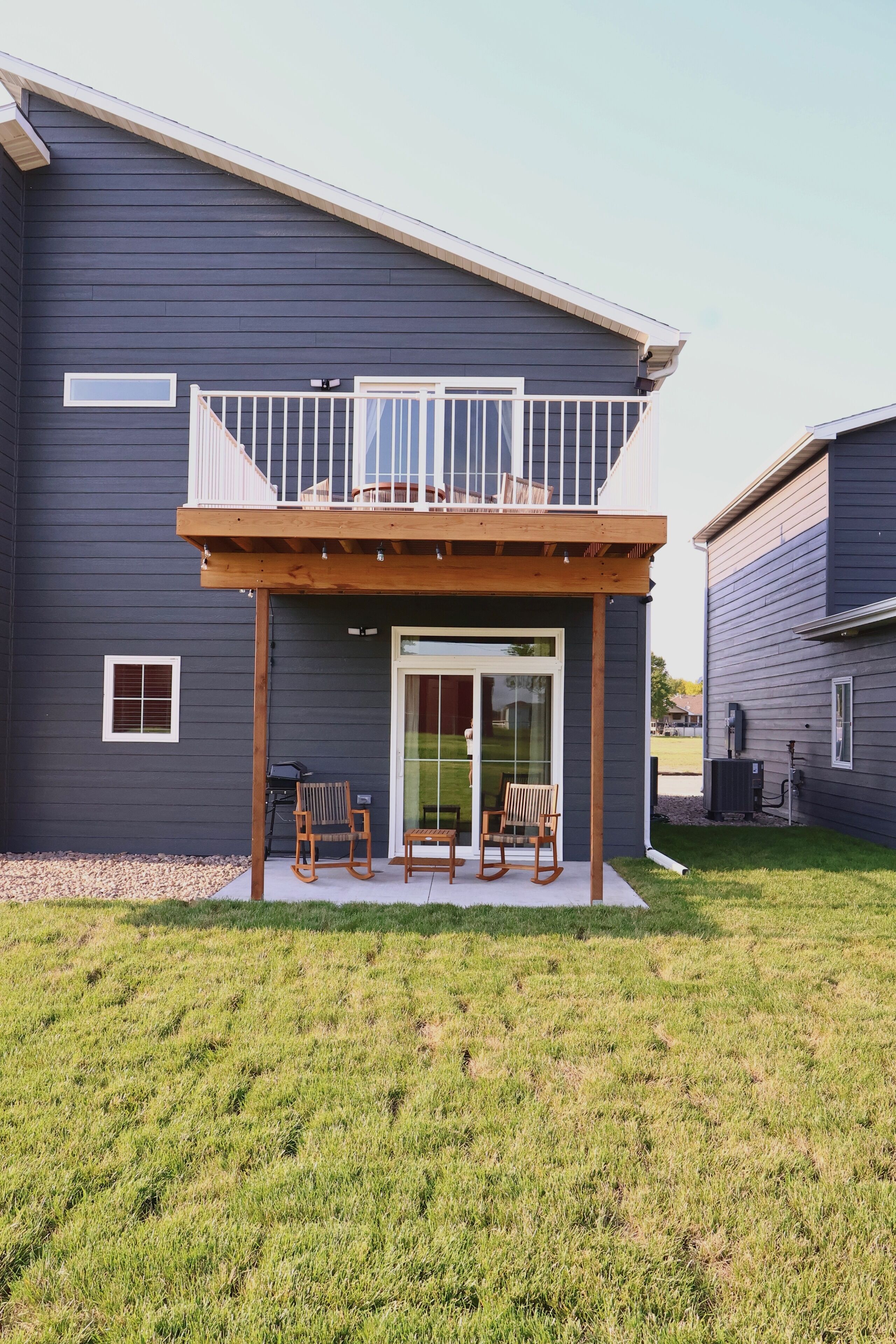 A modern two-story house with a covered patio area, two rocking chairs, and a sliding glass door leading to a backyard.