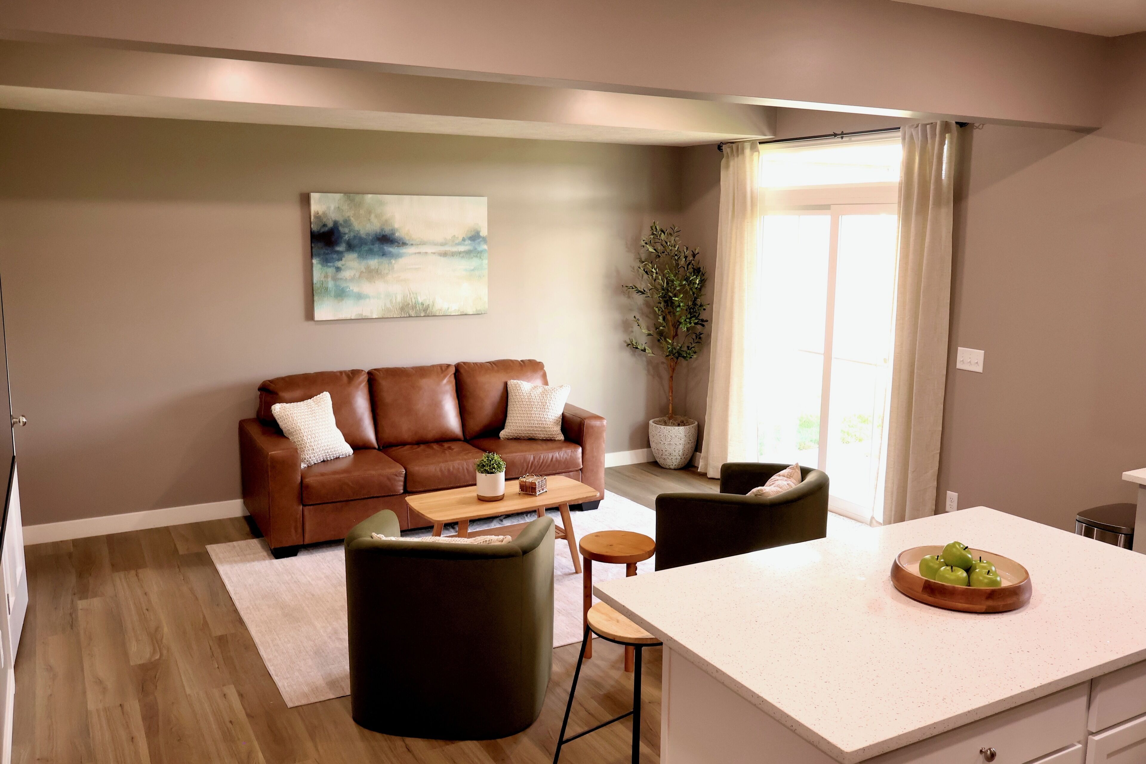 Un salon moderne avec un canapé en cuir marron, une table basse et une petite table ronde accompagnée d’un tabouret. Un tableau orne le mur et une plante en pot se trouve près de la fenêtre.