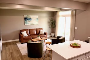 Een moderne woonkamer met een bruine leren bank, een bijzettafeltje en een eethoek met een wit aanrechtblad en een schaal groene appels.