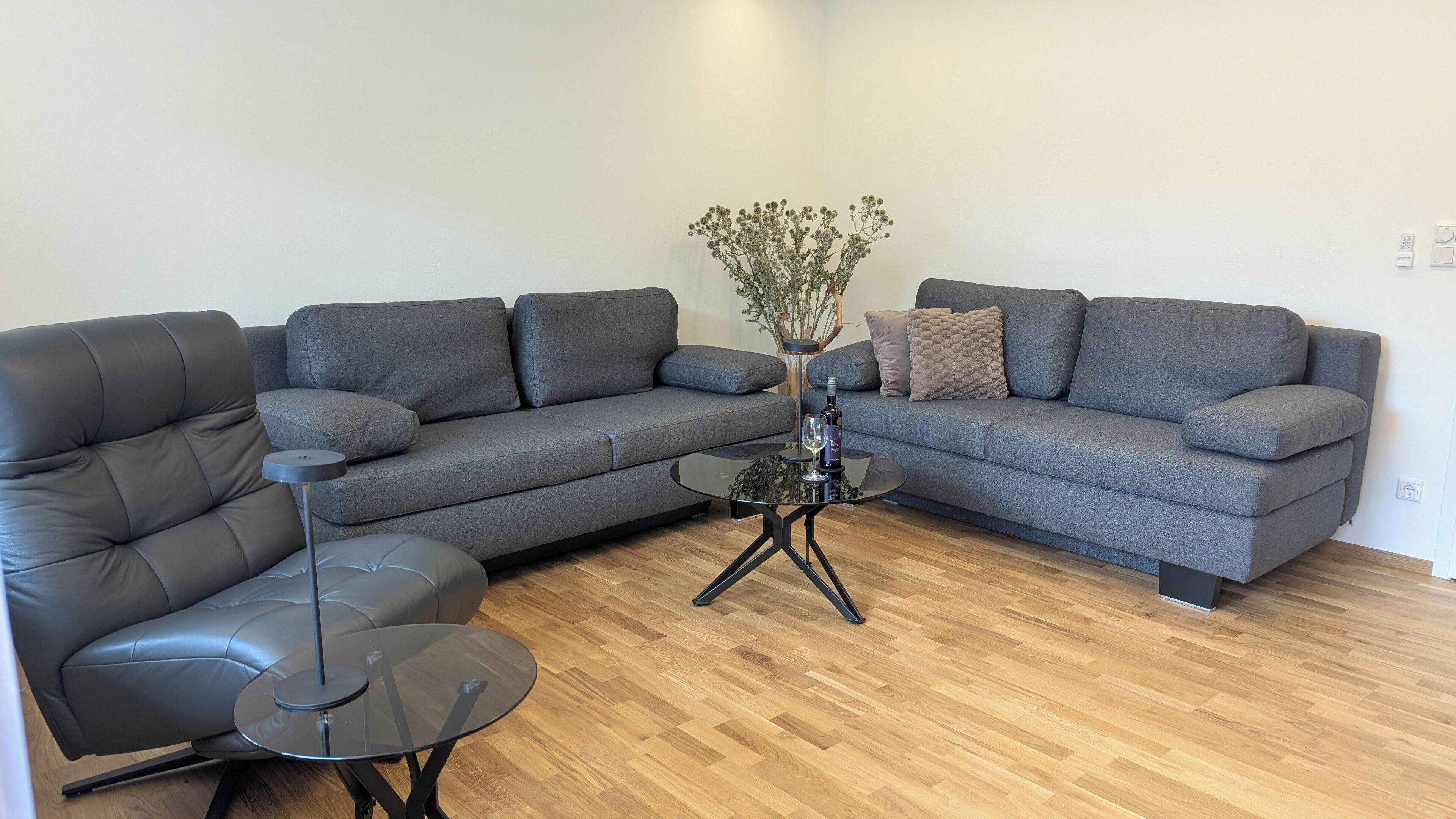 Een moderne woonkamer met een grijze bank, een zwarte leren fauteuil en een glazen salontafel.