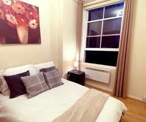 A bedroom with a bed, a nightstand, a radiator, and a window with curtains. - Chelsea 1-Bed Flat on King's Road | Sleeps 3 (London)