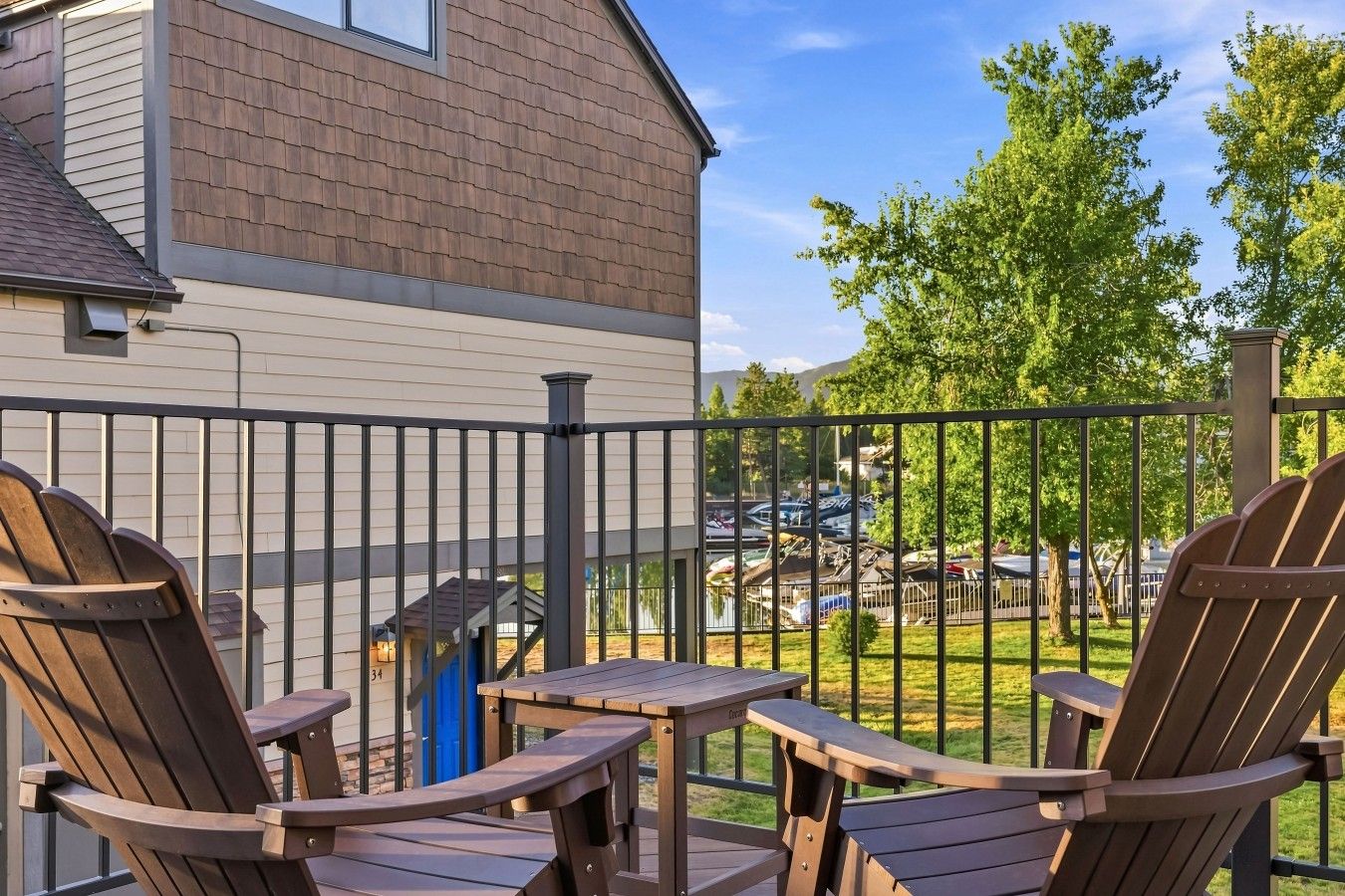 A deck with Adirondack chairs and a small table, overlooking a parking area and trees.