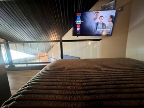 Una habitació amb un llit, una televisió penjada que mostra una pel·lícula, i vistes a l'exterior a través de grans finestres.