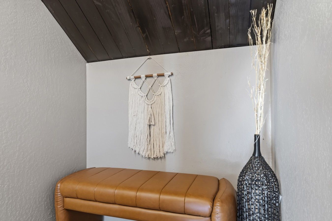 A room with a brown leather bench, a macrame wall hanging, and a vase with dried plants.