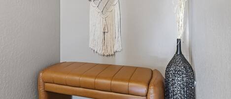 Une pièce avec un banc en cuir marron, une décoration murale en macramé et un vase contenant des plantes séchées.