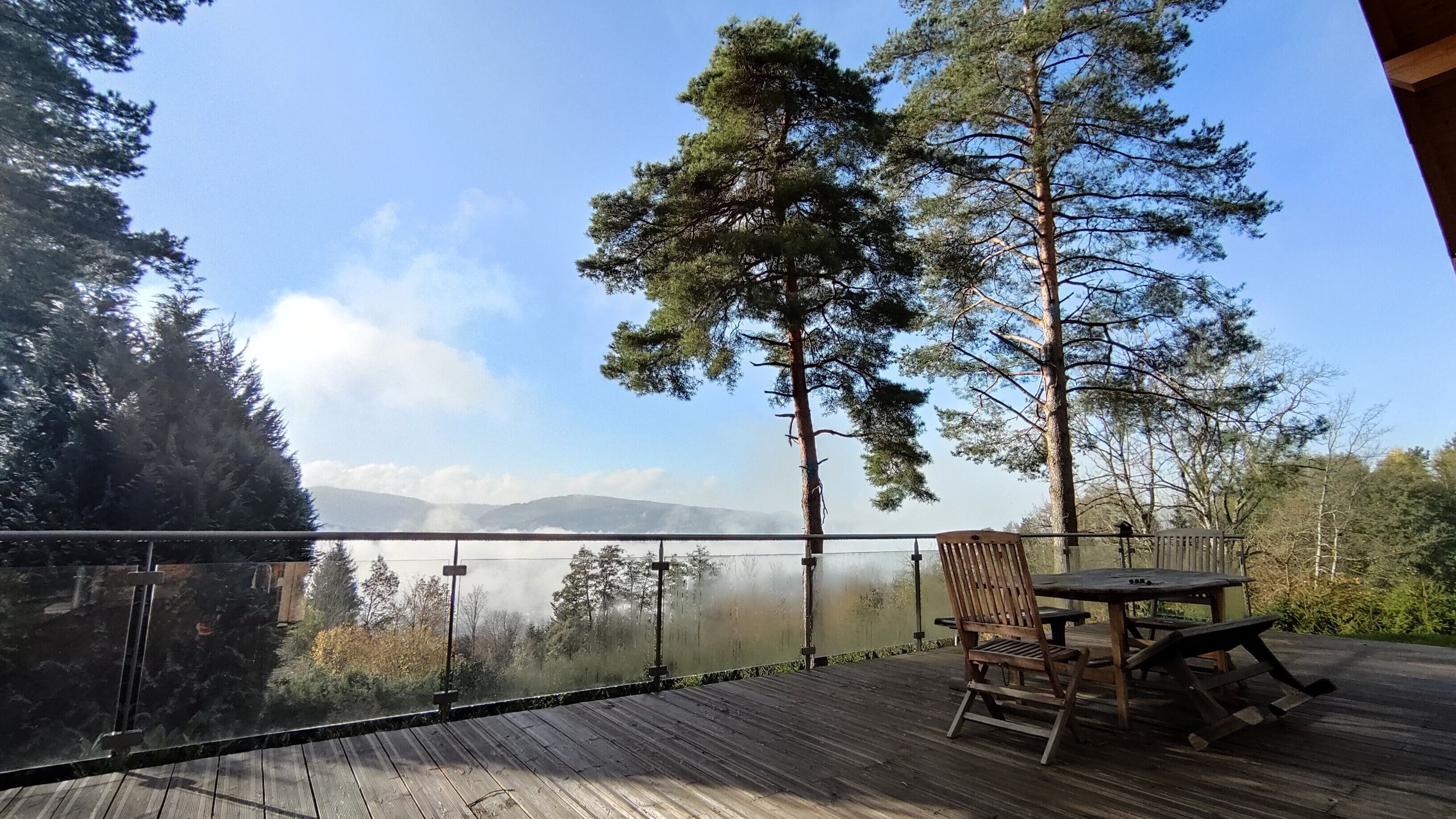 En træterrasse med et bord og stole, der udover et skovområde med bjerge i baggrunden.