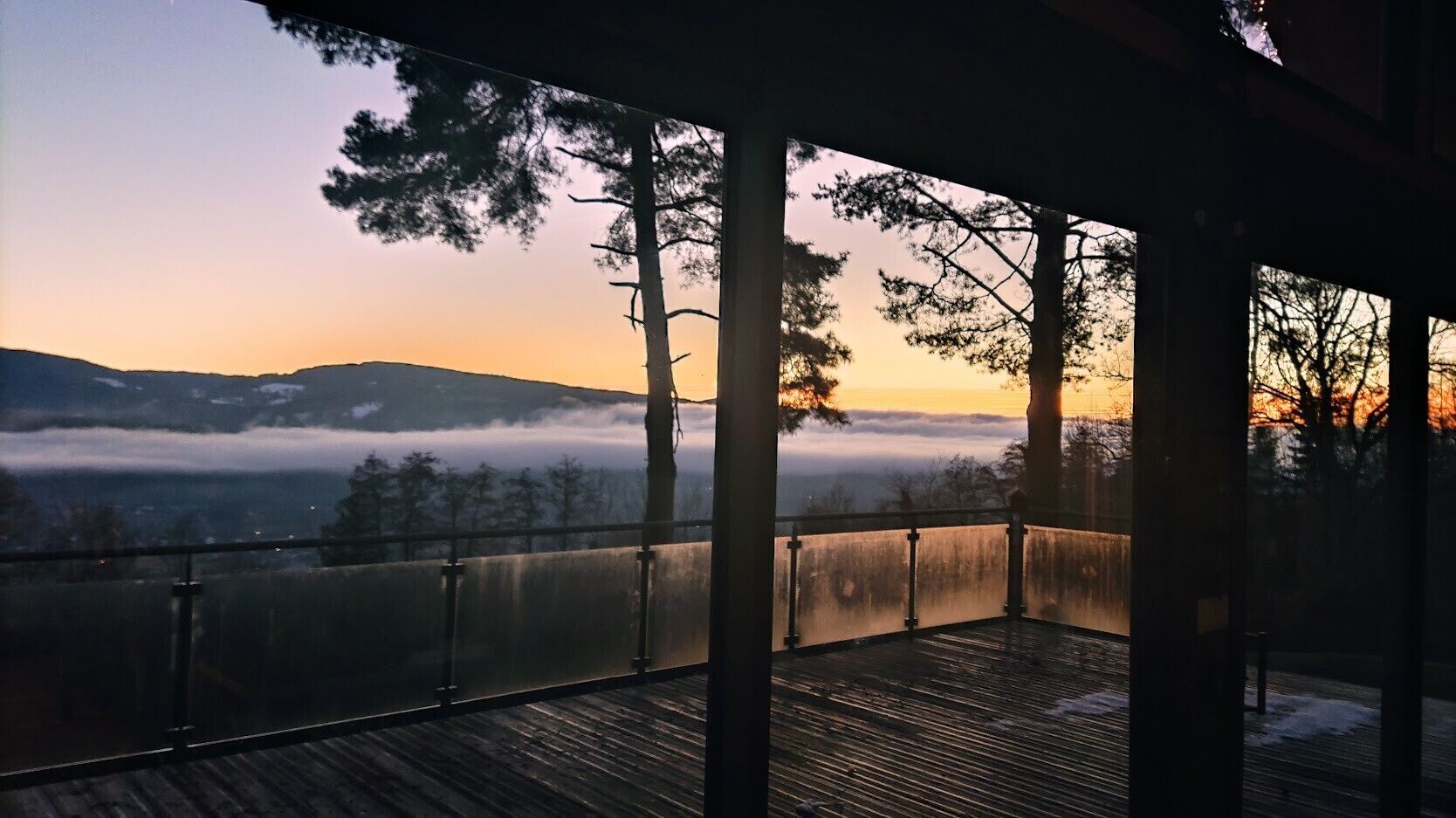 A wooden deck with a glass railing overlooking a mountainous landscape with trees and a sunrise.