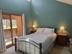 Une chambre avec un lit, deux tables de chevet avec des lampes, une porte-fenêtre coulissante donnant sur un espace extérieur, et une vue sur des arbres et une voiture, visible à travers la porte.